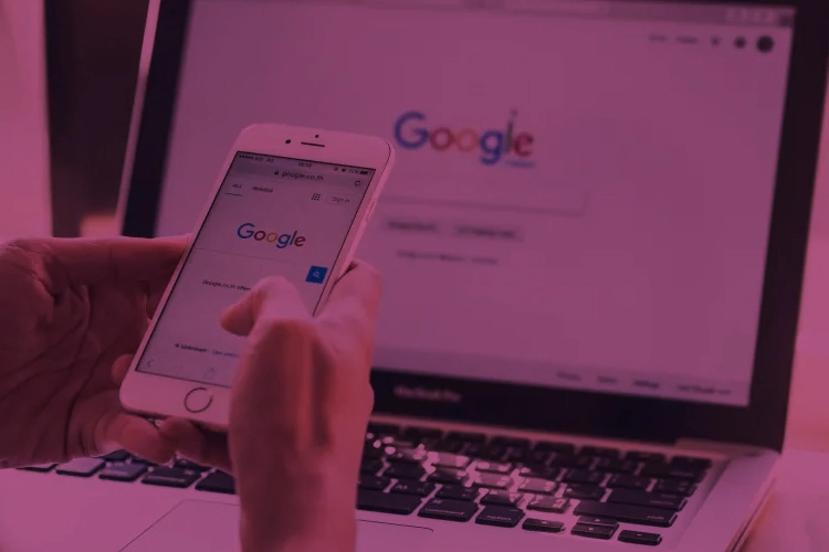 Mãos masculinas segurando um smartphone, aberto na página do Google. Ao fundo há um notebook aberto também na página do Google.