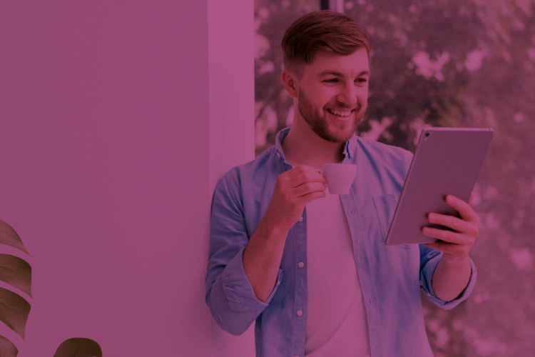 Homem jovem sorrindo e olhando em direção ao tablet, que segura com sua mão esquerda. Com a mão direita, ele segura uma xícara de café. Ele está em um ambiente corporativo, mas usa roupas casuais.